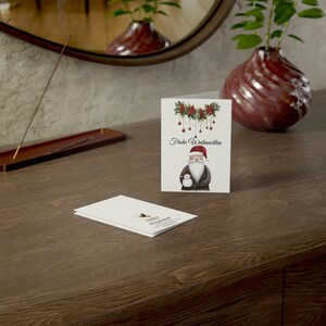 May include: A white card with a watercolor illustration of Santa Claus holding a small snowman. The card has a garland of holly and red ornaments hanging above Santa. The text "Fr&ouml;hliche Weihnachten" is printed on the card.