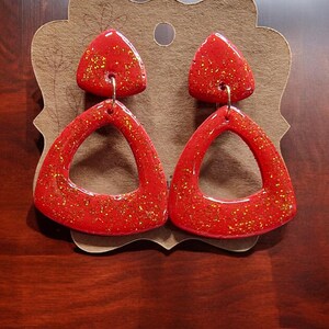 May include: A pair of red glitter triangle earrings with a gold hook. The earrings are made of polymer clay and have a shiny finish.