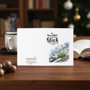 May include: A white greeting card with the German text "Gesundheit und viel Gl&uuml;ck f&uuml;r das neue Jahr" and a watercolor illustration of white flowers on a rocky hillside. A cup of coffee and an open book are in the background.