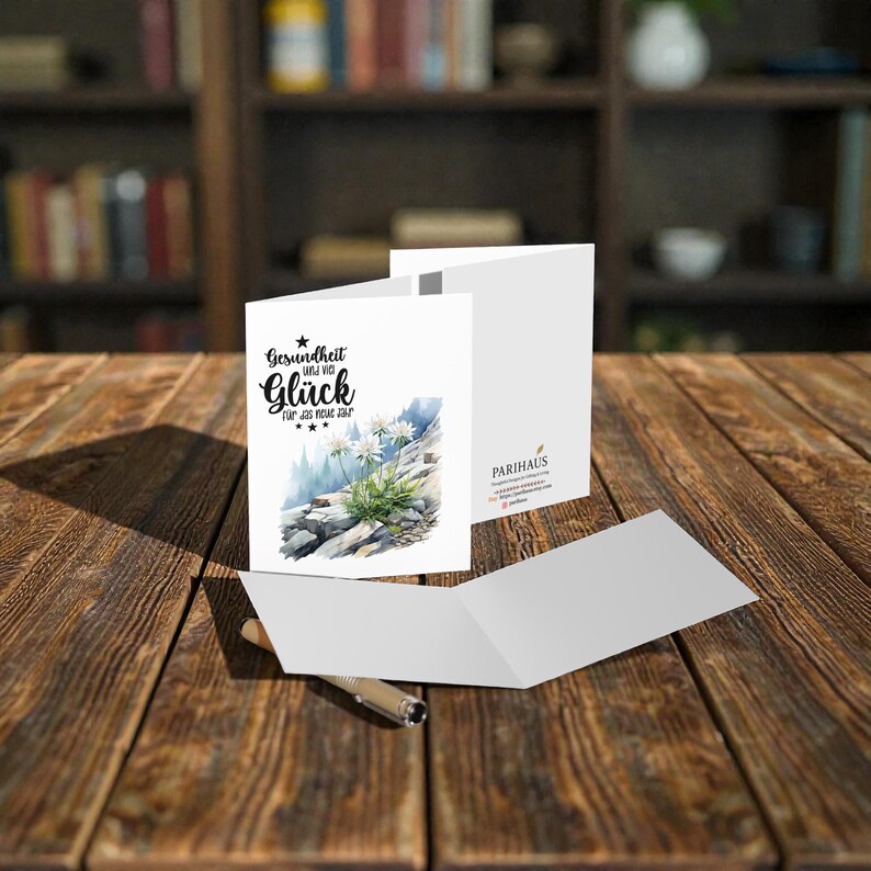 May include: White greeting card with the German text "Gesundheit und viel Gl&uuml;ck f&uuml;r das neue Jahr" and a watercolor illustration of white flowers on a rocky mountain. The card is open, with a pen resting on the wooden surface.