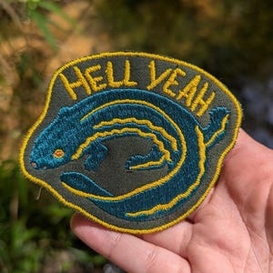 May include: Embroidered patch featuring a stylized blue eel with yellow accents, set against a dark green background. The patch has the words "HELL YEAH" in yellow lettering. The patch is likely designed for clothing or accessories.