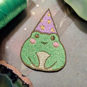 May include: Green embroidered frog wearing a purple and gold star-patterned wizard hat. The frog is sitting with its legs crossed and has a happy expression.