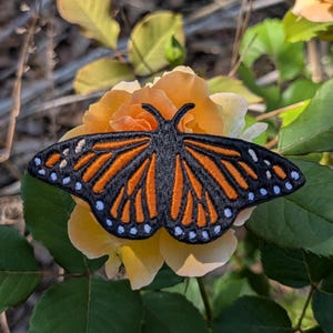 May include: An embroidered butterfly patch with black, orange, and white details, resting on a yellow rose. The butterfly design features intricate wing patterns and white dots along the edges, set against a backdrop of green leaves.