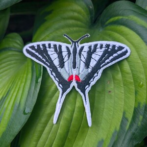 May include: An embroidered patch shaped like a swallowtail butterfly with white wings, black stripes, and two small red spots near the center. The patch features a white border and long, pointed tail-like extensions at the bottom of the wings.