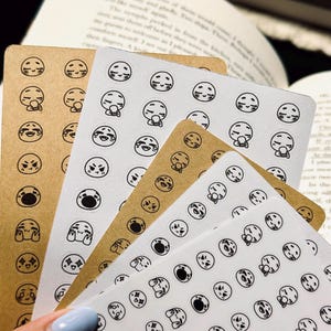 May include: A stack of sticker sheets featuring various emoji expressions in black ink on white and kraft paper. The stickers depict a range of emotions, including joy, sadness, and surprise. The sheets are arranged in a fan shape.