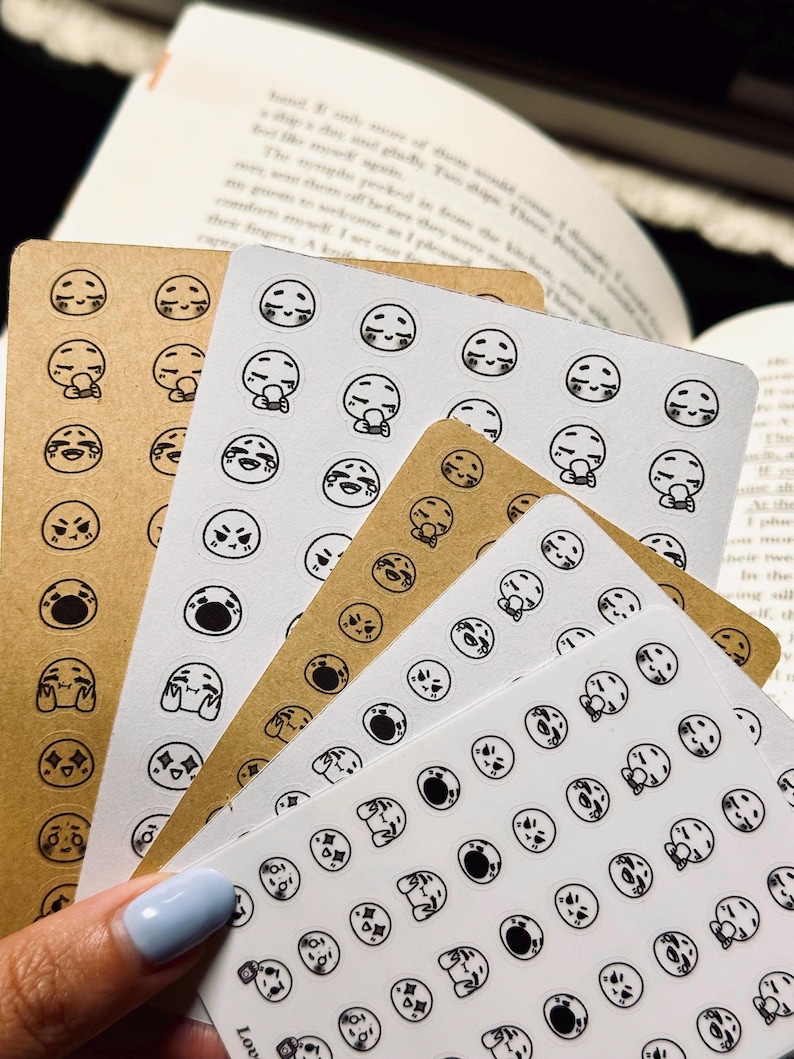May include: A stack of sticker sheets featuring various emoji expressions in black ink on white and kraft paper. The stickers depict a range of emotions, including joy, sadness, and surprise. The sheets are arranged in a fan shape.