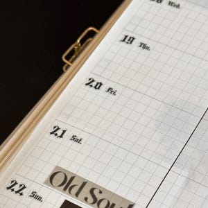 May include: Open planner with grid-lined pages, featuring dates and the words "Old Soul" on a sticker. The planner is held together with a gold-coloured clip. The dates of the week are printed in a classic font.