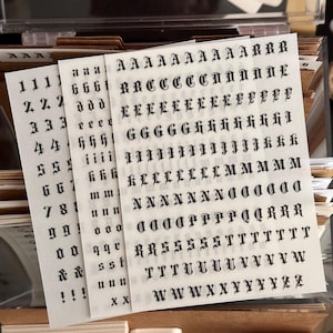 Puede incluir: Tres hojas de pegatinas transparentes con letras, números y símbolos negros de estilo gótico. Las hojas muestran alfabetos en mayúsculas y minúsculas, dígitos del cero al nueve, signos de ampersand y signos de exclamación en filas.