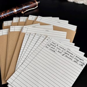 May include: A stack of lined note cards with brown paper envelopes. The cards have the word "Notes" printed at the top and handwritten text. A brown and gold pen rests in the background.
