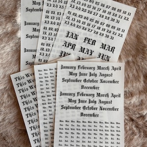 May include: A selection of paper calendar stickers featuring month names, dates, and days of the week printed in black ink. The stickers are displayed on a textured, light brown surface.