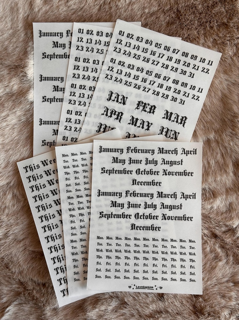May include: A selection of paper calendar stickers featuring month names, dates, and days of the week printed in black ink. The stickers are displayed on a textured, light brown surface.