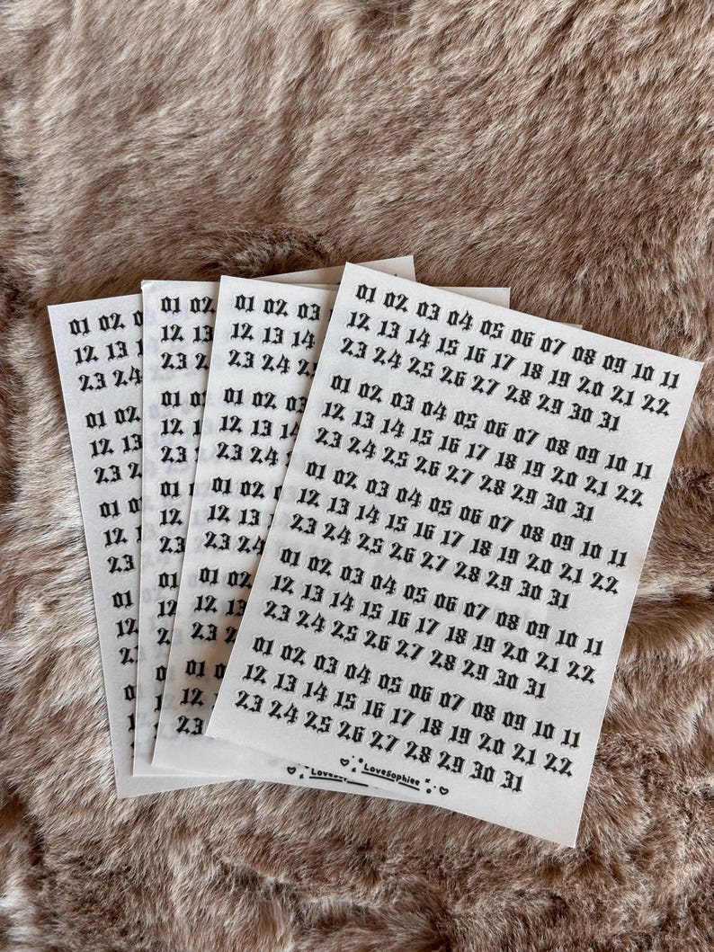 May include: A stack of small white paper sheets with black text, featuring dates like "01 02 03" and "12 13 14", rests on a textured, light brown surface. The sheets are labelled "LoveSophiee".