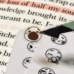 May include: A sheet of small, round stickers featuring various cartoon faces with black outlines and brown accents. A craft knife is cutting a sticker from the sheet, with a book in the background.