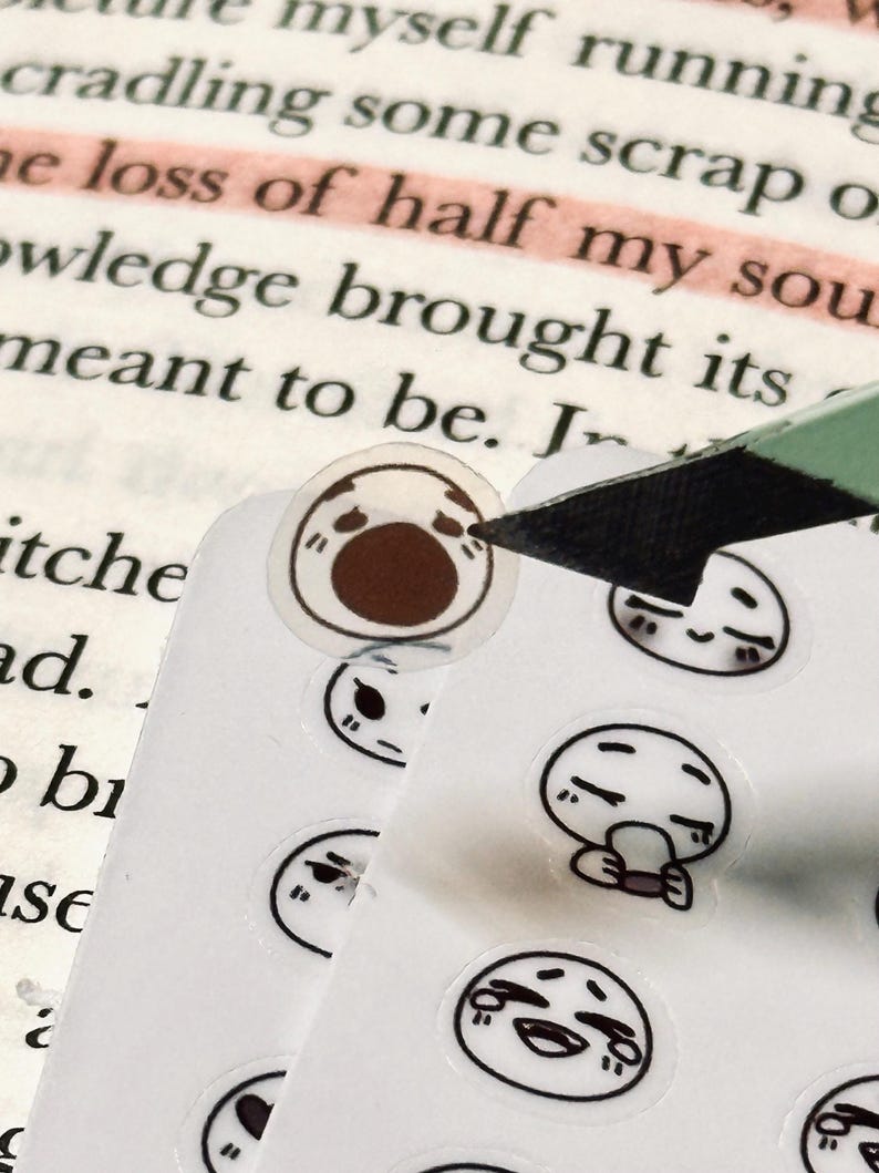 May include: A sheet of small, round stickers featuring various cartoon faces with black outlines and brown accents. A craft knife is cutting a sticker from the sheet, with a book in the background.
