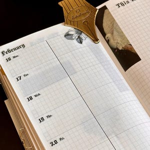May include: A gold-coloured pen nib bookmark sits on an open planner page. The planner shows the week of February 16th, with a grid pattern and the words "This Week". The bookmark is shaped like a pen nib with the word "Esterbrook" on it.