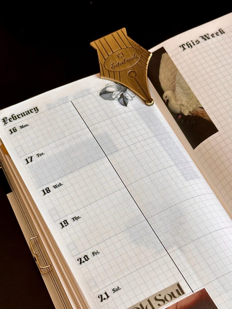 May include: A gold-coloured pen nib bookmark sits on an open planner page. The planner shows the week of February 16th, with a grid pattern and the words "This Week". The bookmark is shaped like a pen nib with the word "Esterbrook" on it.