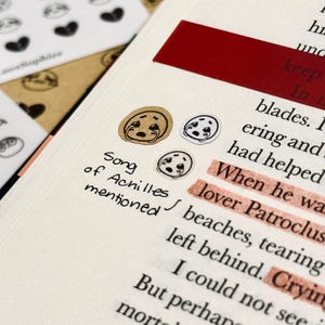 May include: Close-up of a journal page with text and decorative stickers. The stickers feature a crying face design. The text reads "Song of Achilles mentioned" with highlighted words. A red rectangular sticker is also visible.