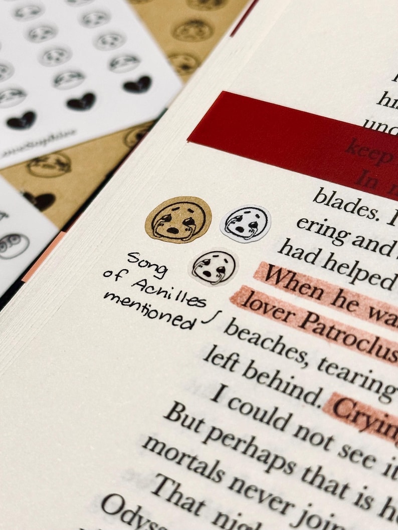 May include: Close-up of a journal page with text and decorative stickers. The stickers feature a crying face design. The text reads "Song of Achilles mentioned" with highlighted words. A red rectangular sticker is also visible.