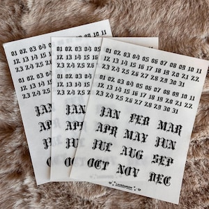 May include: Three sheets of white paper with black text. Each sheet has the months of the year and dates from 01 to 31. The text is in a gothic font. The background is a brown, textured surface.