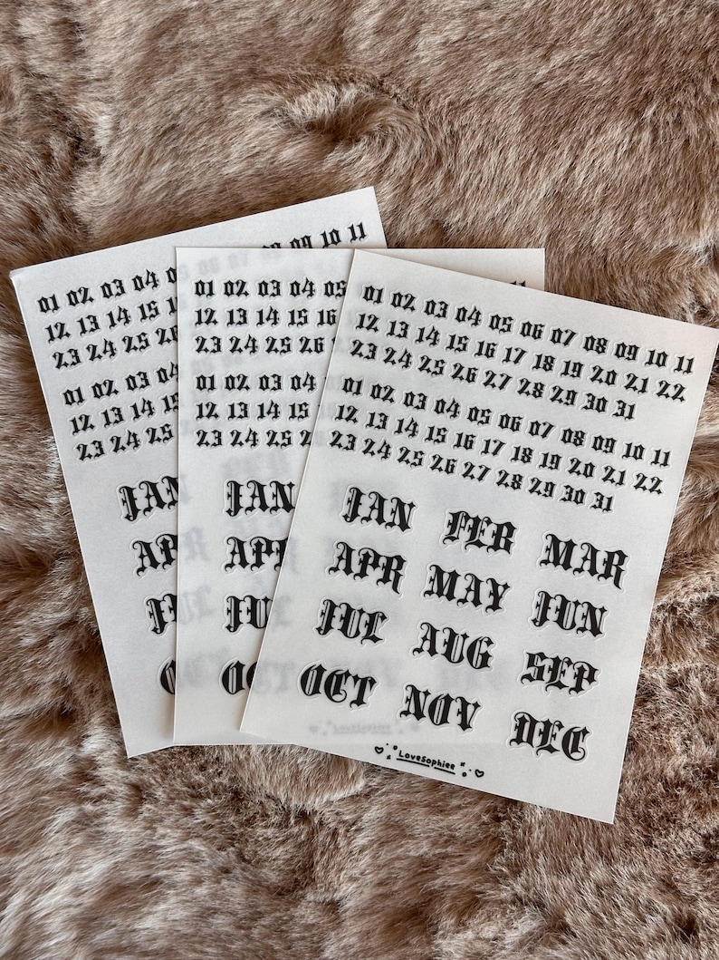 May include: Three sheets of white paper with black text. Each sheet has the months of the year and dates from 01 to 31. The text is in a gothic font. The background is a brown, textured surface.