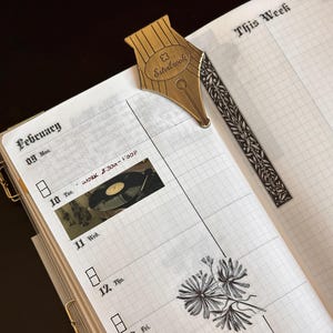 May include: An open planner showing the February section, with a gold-coloured pen nib bookmark. The planner has a grid layout with handwritten text and stickers. The bookmark is shaped like a pen nib with the word "Esterbrook" on it.