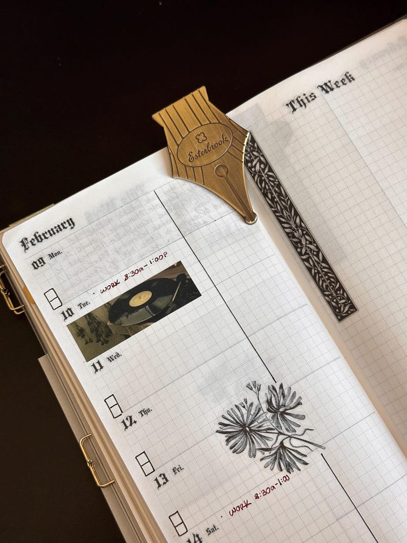 May include: An open planner showing the February section, with a gold-coloured pen nib bookmark. The planner has a grid layout with handwritten text and stickers. The bookmark is shaped like a pen nib with the word "Esterbrook" on it.