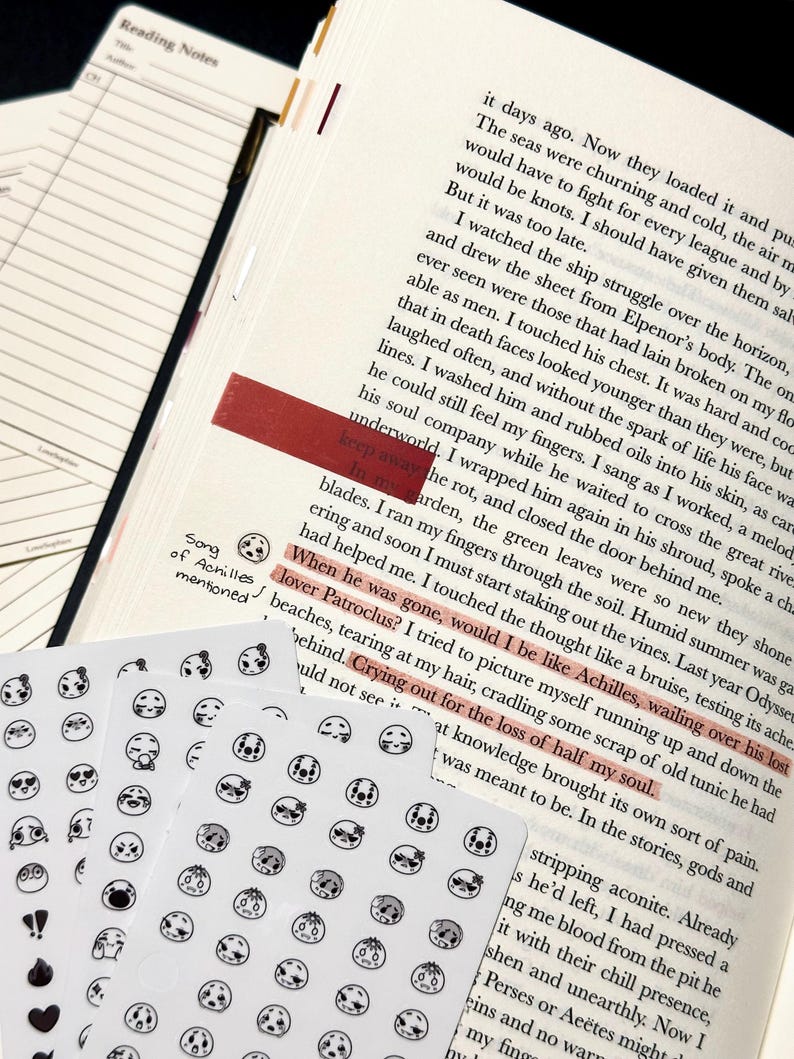 May include: A stack of white sticker sheets featuring various small, round faces with different expressions. The stickers are placed near an open book with highlighted text and a red bookmark, suggesting a reading or journaling theme.