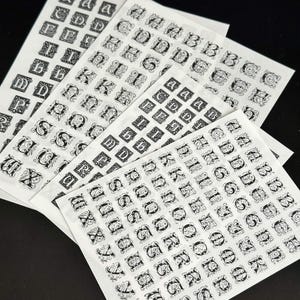 Könnte beinhalten: Mehrere Bögen mit Aufklebern, die schwarze, verzierte Buchstaben im gotischen Stil als Groß- und Kleinbuchstaben zeigen. Jeder Buchstabe ist in einem dekorativen quadratischen Rahmen mit komplexen Schnörkeldetails eingeschlossen und alphabetisch angeordnet.