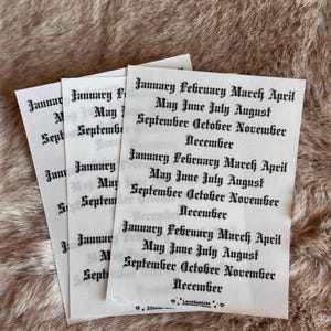 May include: Three white paper sheets with the months of the year printed in a gothic font. The text includes "January February March April May June July August September October November December". The sheets are on a brown, textured surface.