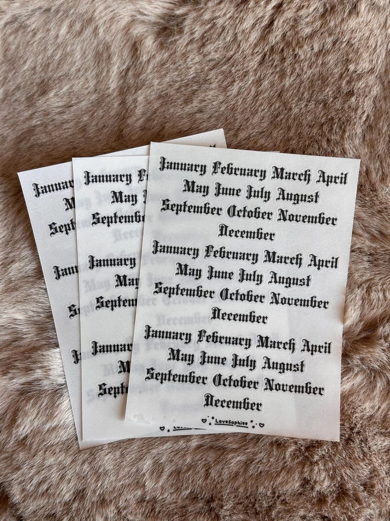 May include: Three white paper sheets with the months of the year printed in a gothic font. The text includes "January February March April May June July August September October November December". The sheets are on a brown, textured surface.