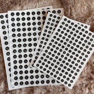 Könnte beinhalten: Blätter mit schwarz-weißen Alphabetaufklebern. Jeder Aufkleber zeigt einen Buchstaben des Alphabets in einem schwarzen Kreis. Die Aufkleber sind auf weißem Papier angeordnet und auf einer strukturierten, braunen Oberfläche dargestellt.