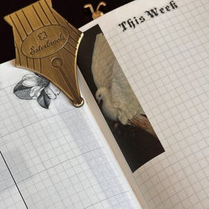 May include: A brass pen nib bookmark, featuring the Esterbrook logo, sits on a grid-lined notebook. A dove image and the words "This Week" are also visible. The notebook is open, revealing the interior pages.