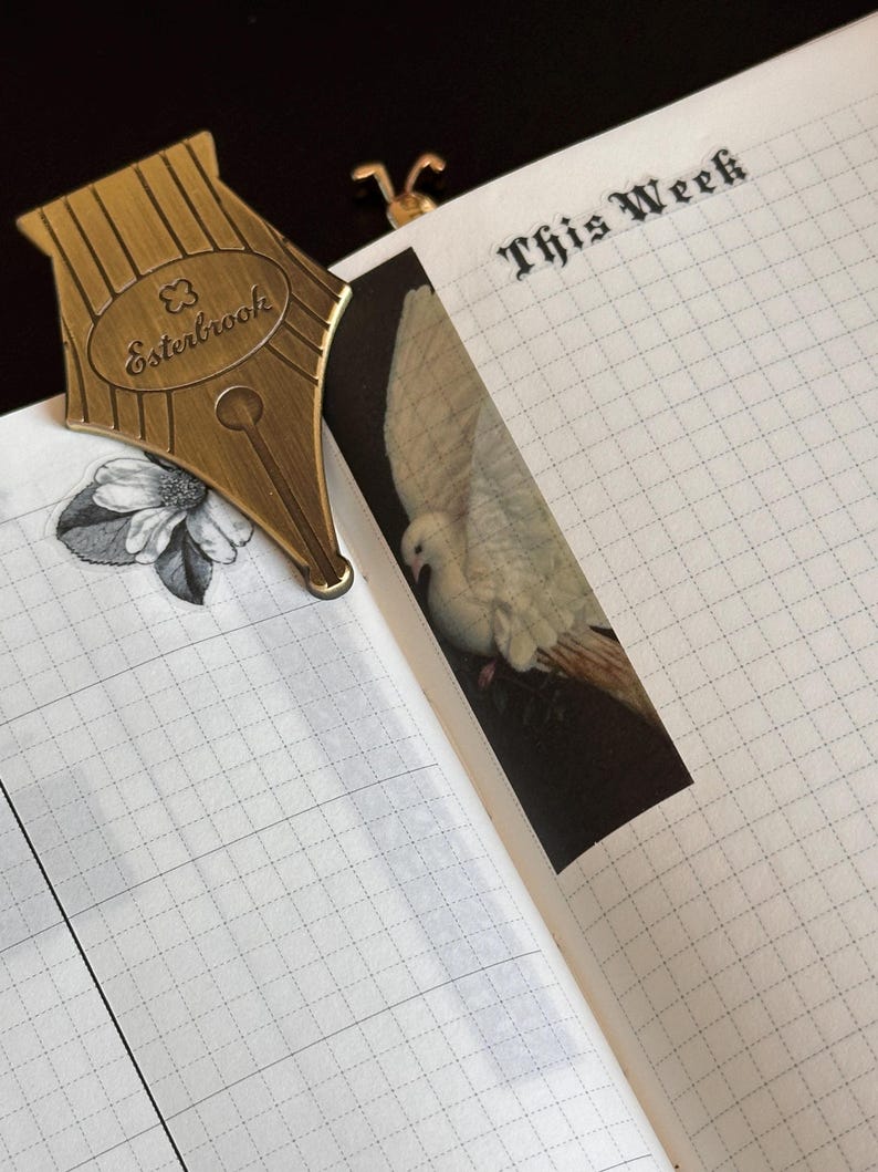 May include: A brass pen nib bookmark, featuring the Esterbrook logo, sits on a grid-lined notebook. A dove image and the words "This Week" are also visible. The notebook is open, revealing the interior pages.
