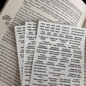 May include: A stack of small, translucent stickers with handwritten text, including phrases like "Omg!" and "Plot twist!" are placed on an open book. The stickers have a handwritten aesthetic, and the book's pages are visible in the background.