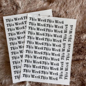 May include: Two white sticker sheets featuring the phrase "This Week" in a gothic font. The stickers are placed on a textured, brown surface. The dark text contrasts with the light background, creating a visually appealing design.