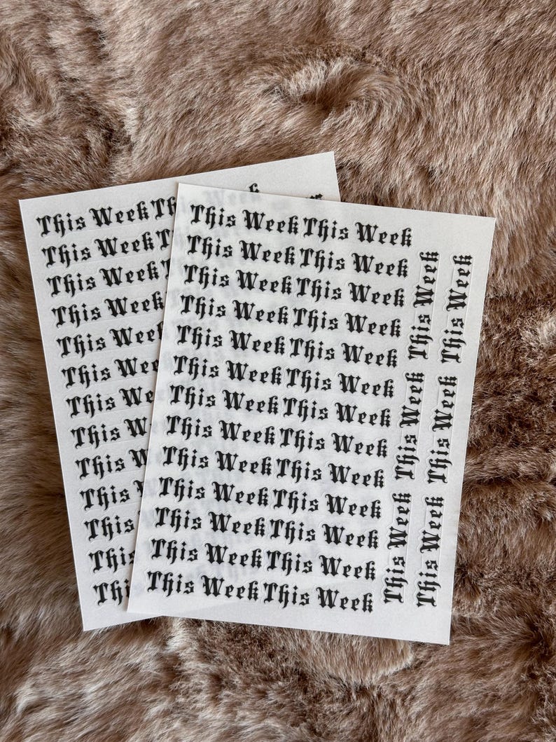 May include: Two white sticker sheets featuring the phrase "This Week" in a gothic font. The stickers are placed on a textured, brown surface. The dark text contrasts with the light background, creating a visually appealing design.
