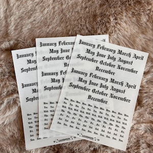 May include: Three white paper sheets with black text, featuring month and day names in a gothic font. The sheets are arranged on a textured, light brown surface. The text includes months like January, May, and September.