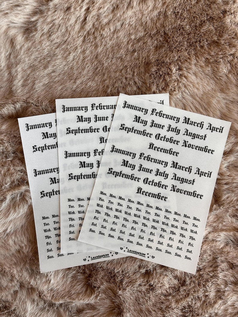 May include: Three white paper sheets with black text, featuring month and day names in a gothic font. The sheets are arranged on a textured, light brown surface. The text includes months like January, May, and September.