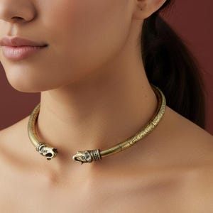 May include: A gold-toned choker necklace with an intricate design. The necklace has a tubular form, adorned with detailed carvings and animal head motifs at each end. The background is a deep red.