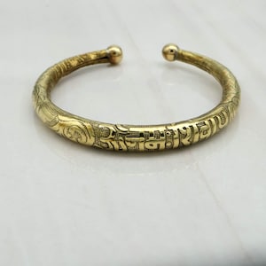 Puede incluir: Una pulsera de brazalete dorada con intrincados patrones grabados y texto. El diseño abierto presenta terminales redondeados en forma de bola. La superficie de la pulsera está adornada con elementos florales y de escritura detallados, creando una estética vintage.