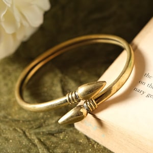May include: A gold-coloured metal bracelet with a distinctive design. The bracelet has two pointed ends that meet in the middle. The bracelet is resting on a green surface.