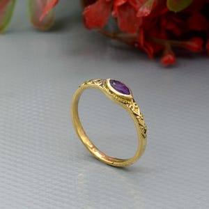 May include: A gold ring with an oval, purple gemstone. The ring has an ornate design along the band and around the gemstone. The ring is set against a gray background with red flowers in the background.