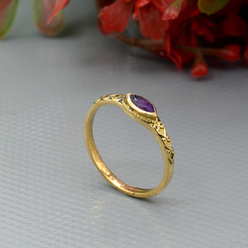 May include: A gold ring with an oval, purple gemstone. The ring has an ornate design along the band and around the gemstone. The ring is set against a gray background with red flowers in the background.