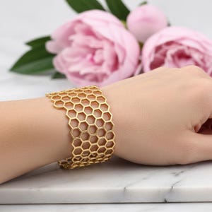 Könnte beinhalten: Ein goldfarbenes Armband mit Wabenmuster. Das Armband wird an einem Handgelenk getragen, mit rosa Blumen im Hintergrund. Das offene Design des Armbands besteht aus vielen sechseckigen Formen.