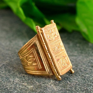 May include: A gold ring with an elaborate rectangular design. The ring has detailed geometric patterns. The background is a blurred green and grey.