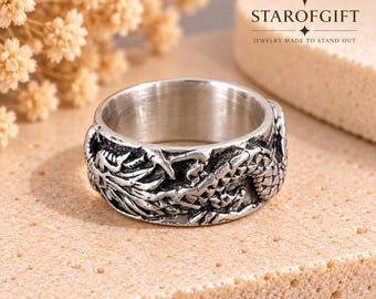 Può includere: Un anello in metallo color argento con un intricato design a drago in rilievo che avvolge l'intera fascia. La superficie presenta una finitura ossidata scura per evidenziare i dettagli delle squame e degli artigli. Il testo STAROFGIFT è visibile.