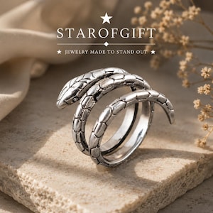 Può includere: Un anello in metallo color argento a forma di serpente avvolto con dettagli a scaglie testurizzate. L'anello presenta un design avvolgente. Sopra l'anello, è visualizzato il testo STAROFGIFT con la frase JEWELRY MADE TO STAND OUT sotto.