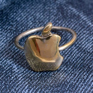 May include: A silver ring with a gold-colored apple-shaped charm. The apple charm has a stem and a bite taken out of it. The ring is set against a blue denim background.
