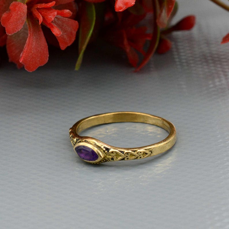 May include: A gold-colored ring with an oval, purple gemstone. The ring has an ornate design on the band, with a filigree pattern near the gemstone. The ring is set against a gray background with red flowers.