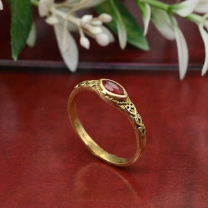 Puede incluir: Un anillo de oro con una piedra preciosa roja. El anillo tiene un diseño ornamentado con detalles de filigrana. La piedra preciosa, de forma ovalada, está engastada en un bisel de oro. El anillo se muestra sobre una superficie roja.